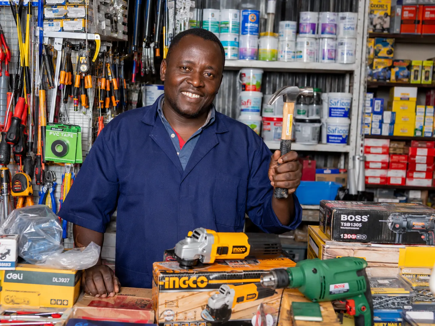 Smiling hardware shop owner in a navy work coat holding a hammer behind a counter stacked with power tools